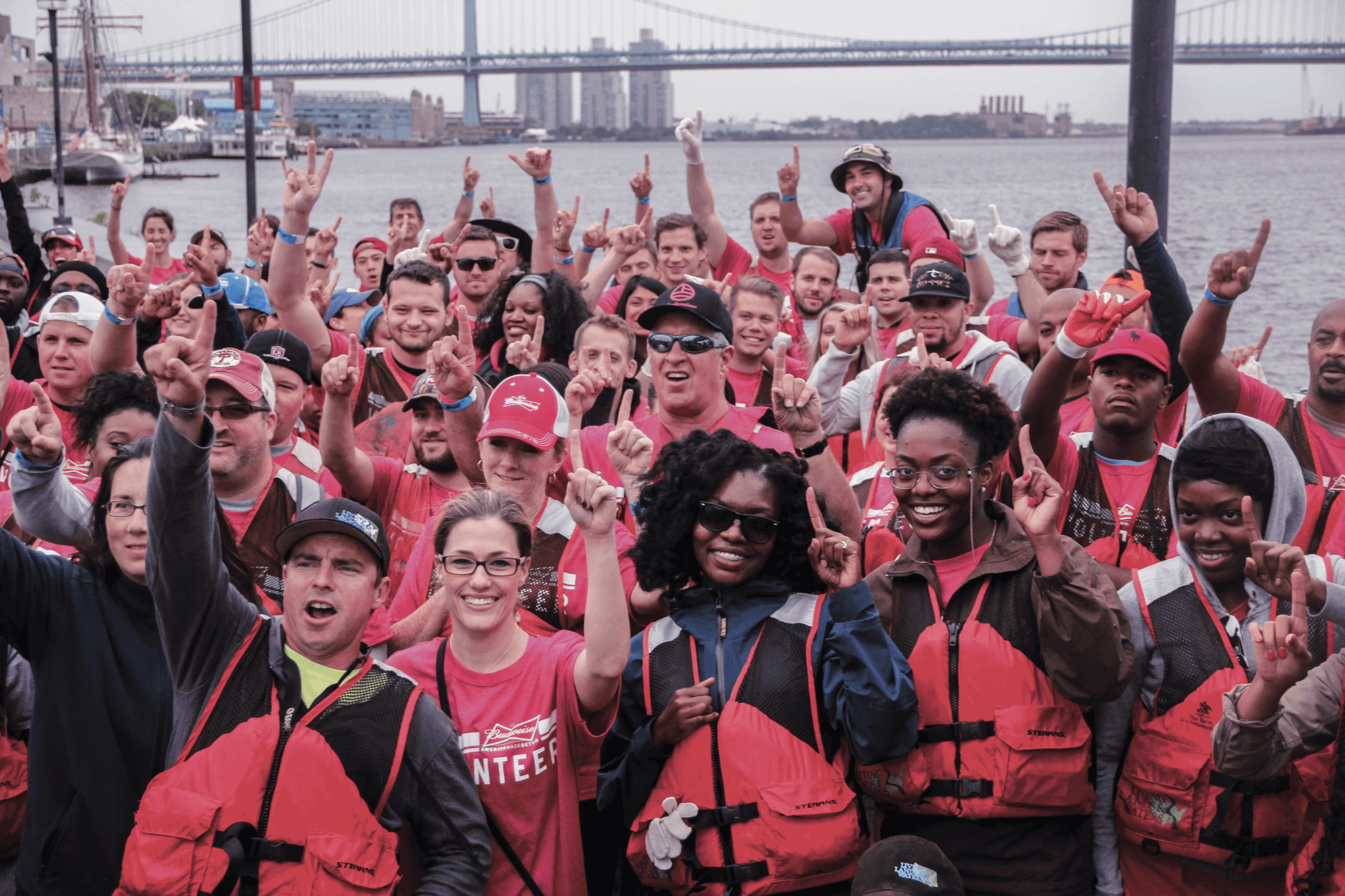 A large group of people stands together cheering and holding up their hands, in front of a river in Philadelphia.