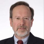 Headshot of Richard Roos-Collins from the shoulders up, wearing frameless glasses and a red tie, looking into the camera on a white background.