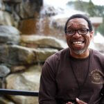 Darryl Haddock standing and smiling in front of a rock wall with trees in the background.