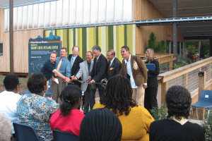 A row of people stands before a seated audience outside a new building for a ribbon cutting ceremony.