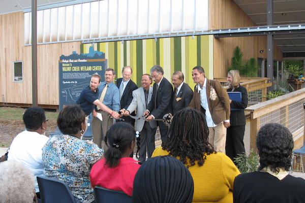 A row of people stands before a seated audience outside a new building for a ribbon cutting ceremony.