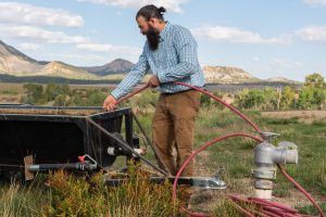 Rancher Ben Wolcott filling a black bin with water using a red hose, with mountains and clouds in the background.