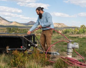 Rancher Ben Wolcott filling a black bin with water using a red hose, with mountains and clouds in the background.