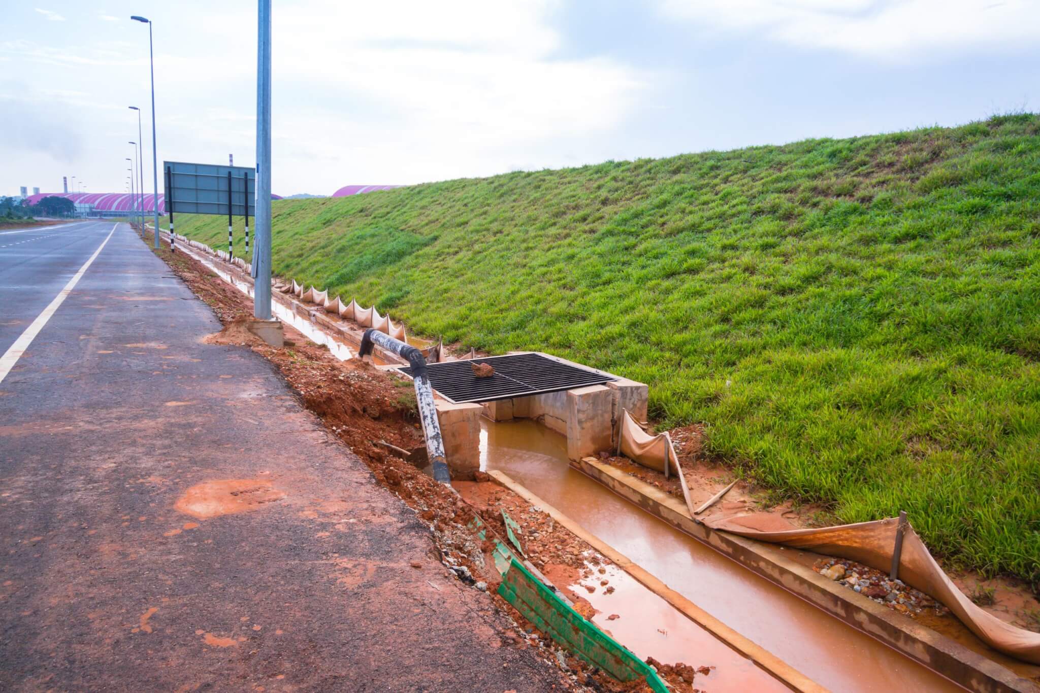 A roadway on the left is dirtied by muddy water that runs parallel to the road. To the right, a sloping grass strip has a fenced barrier next to the water to prevent erosion, parts of the barriers are broken.