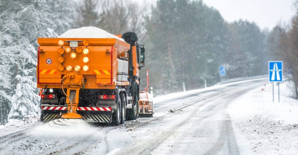 A large orange truck disperses salt onto a snow covered highway with snow covered trees in the background.