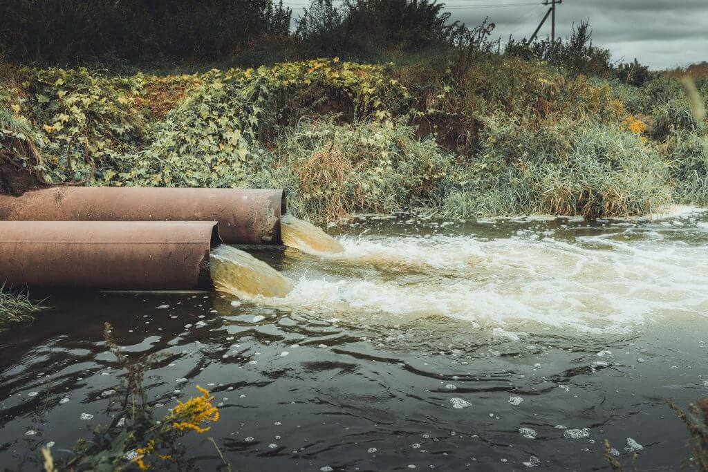 Two brown metal combined sewage overflow pipes release water next to a hillside covered in vegetation.