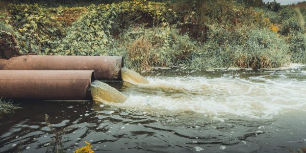 Two brown metal combined sewage overflow pipes release water next to a hillside covered in vegetation.