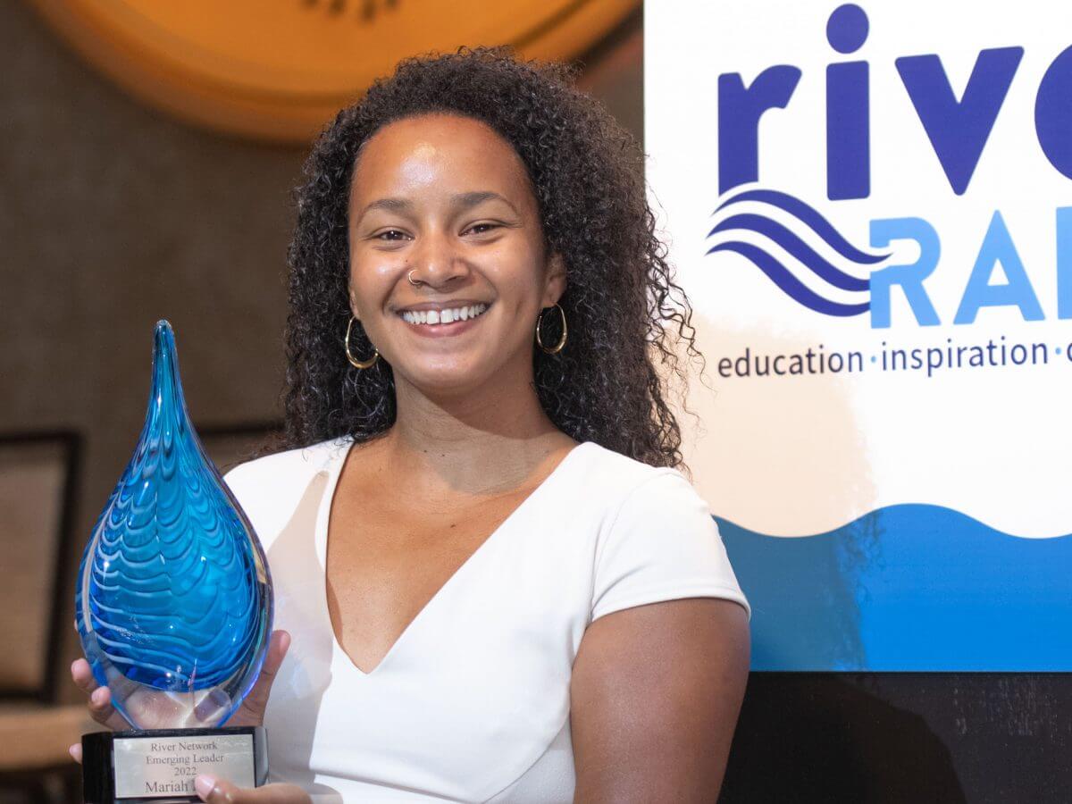 Mariah Davis (she/her) standing in front of the podium on the River Rally stage holding her Emerging Leader award.