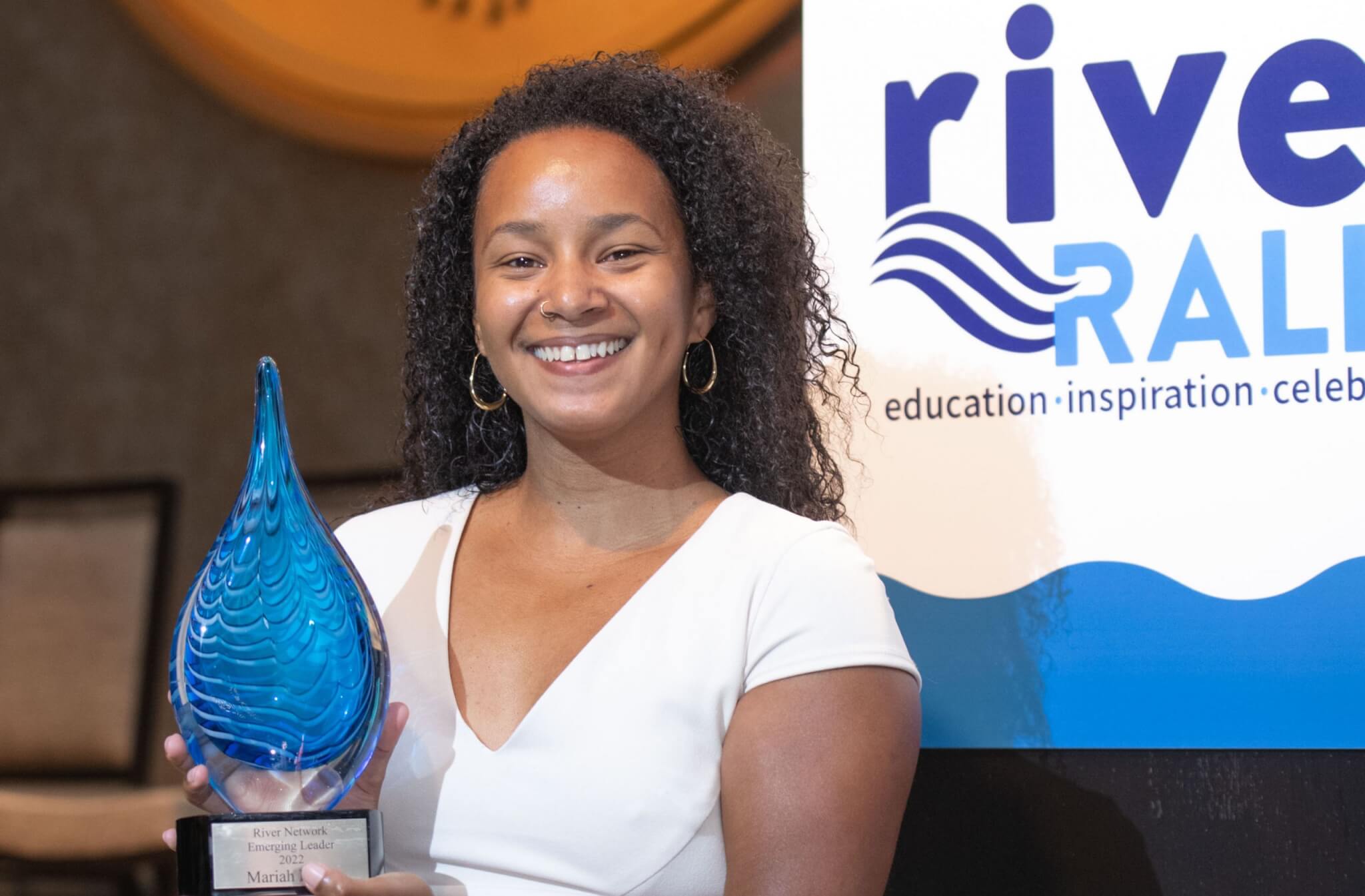 Mariah Davis (she/her) standing in front of the podium on the River Rally stage holding her Emerging Leader award.
