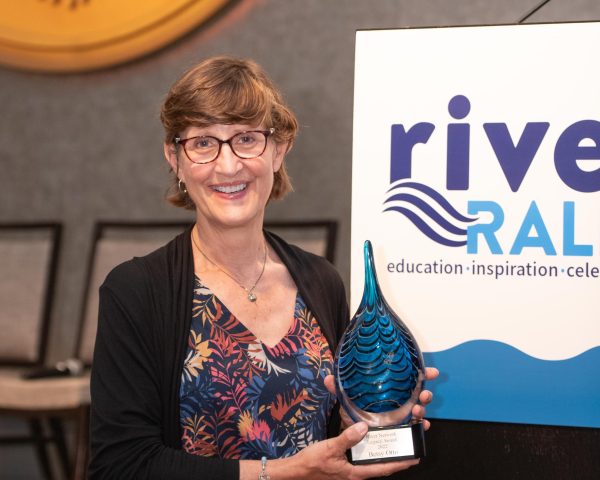 Betsy Otto (she/her) standing in front of the podium on the River Rally stage holding her Legacy award.