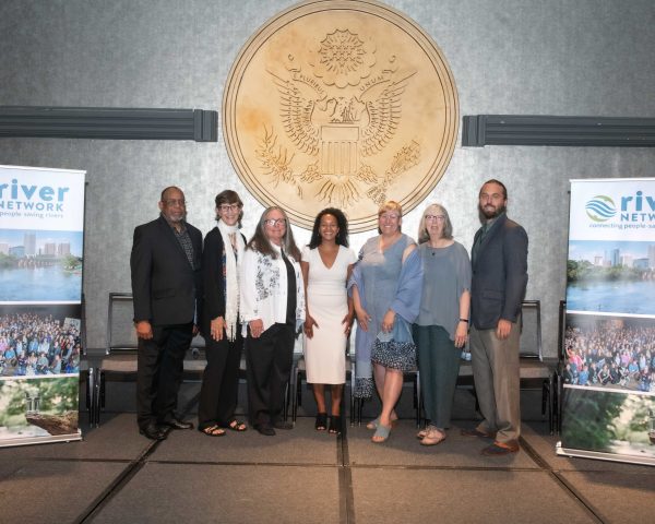 Seven awardees standing in a line the River Rally stage. Awardees are, from left to right, Arthur Johnson, Betsy Otto, Christina McVie, Mariah Davis, Melanie Ariens, Ann-Marie Mitroff, and Trey Sherard.