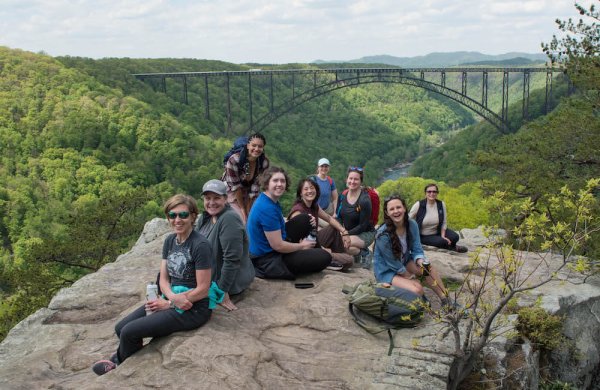 River Network staff sitting on a large rock with a majestic bridge, trees, and river in the background. Photo taken during a hike in West Virginia.