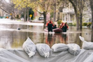 Five white sandbags rest on a makeshift dam, holding back floodwater along a city street lined with trees. Some people are floating in a raft in the background.