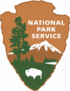 Brown arrowhead logo, point down. At top right, white text, National Park Service. At left, a tall tree. At bottom, a white bison stands on a green field ending in a distant tree line, a white lake at right. A snow-capped mountain towers behind.