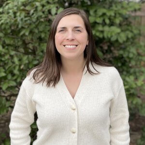 Headshot of Stephanie Heidbreder. She's wearing a white button down cardigan sweater, standing in front of a leafy background.